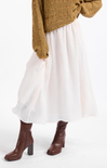 A person wearing a flowing white midi skirt, a knitted mustard-brown sweater, and brown heeled ankle boots against a plain white studio background.