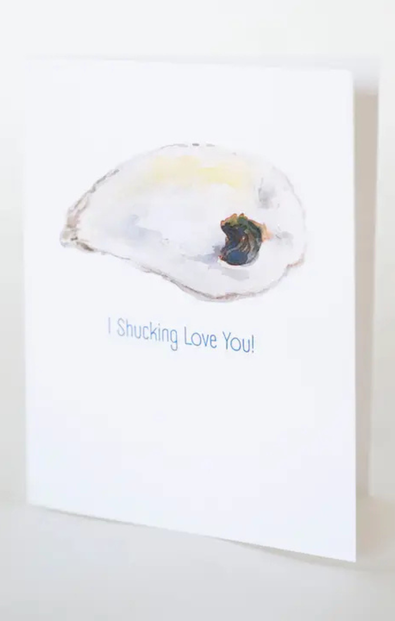 A white greeting card featuring a watercolor illustration of an oyster shell with the text 'I Shucking Love You!' printed below in blue.