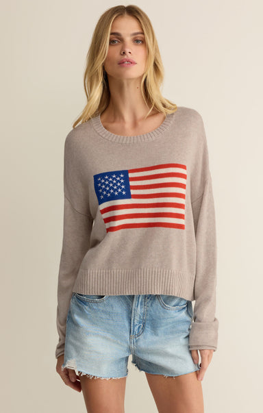 Woman wearing a beige sweater with an American flag design and denim shorts on a plain background