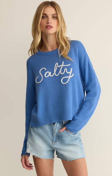 Woman wearing a blue sweater with 'Salty' text and denim shorts on a beige background