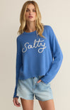 Woman wearing a blue sweater with 'Salty' text and denim shorts on a beige background