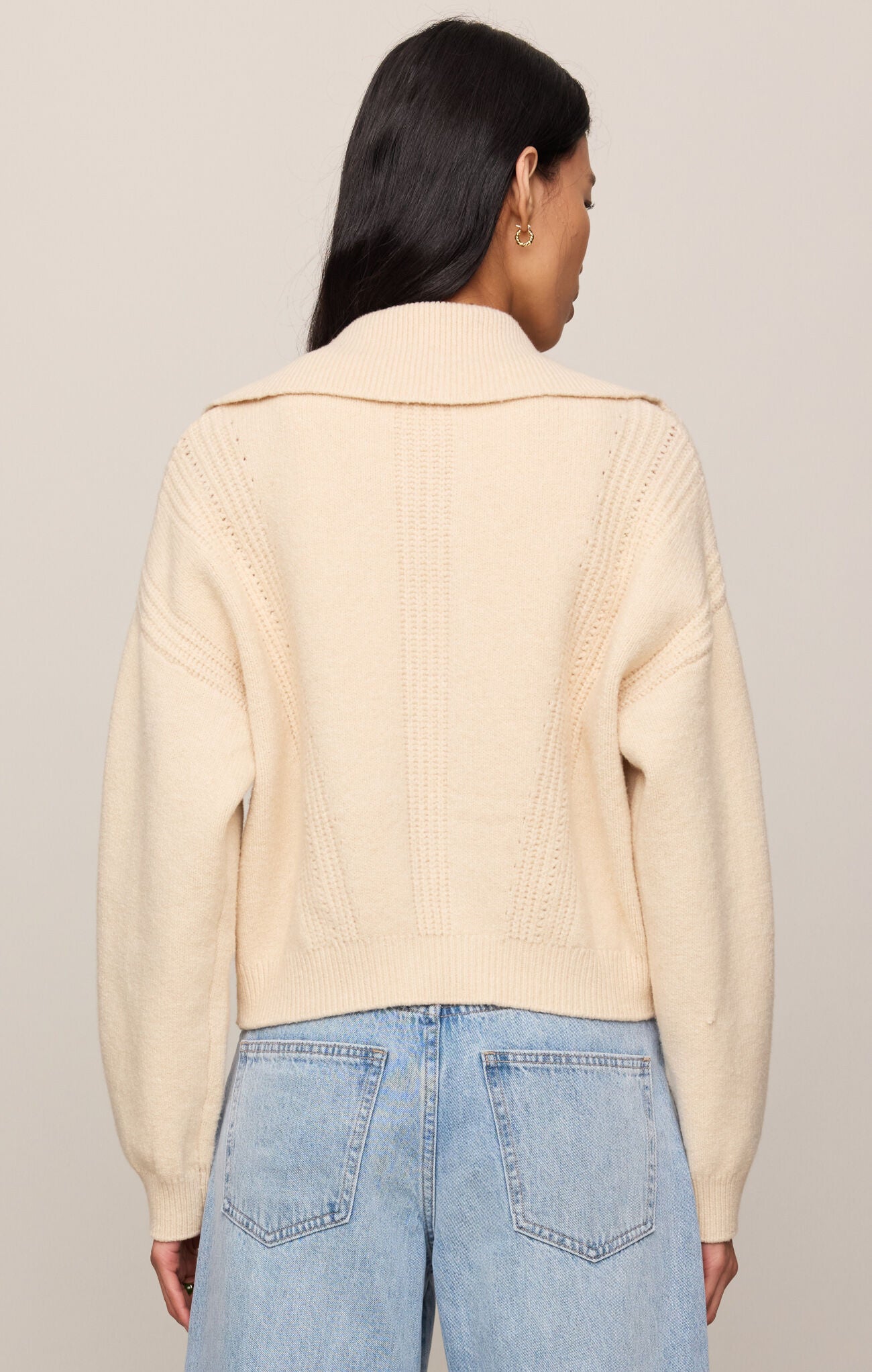 Person wearing a beige ribbed sweater and blue jeans against a neutral background