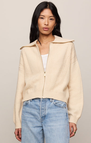 Woman wearing a beige cardigan over a white top and blue jeans on a plain background