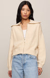 Woman wearing a beige cardigan over a white top and blue jeans on a plain background