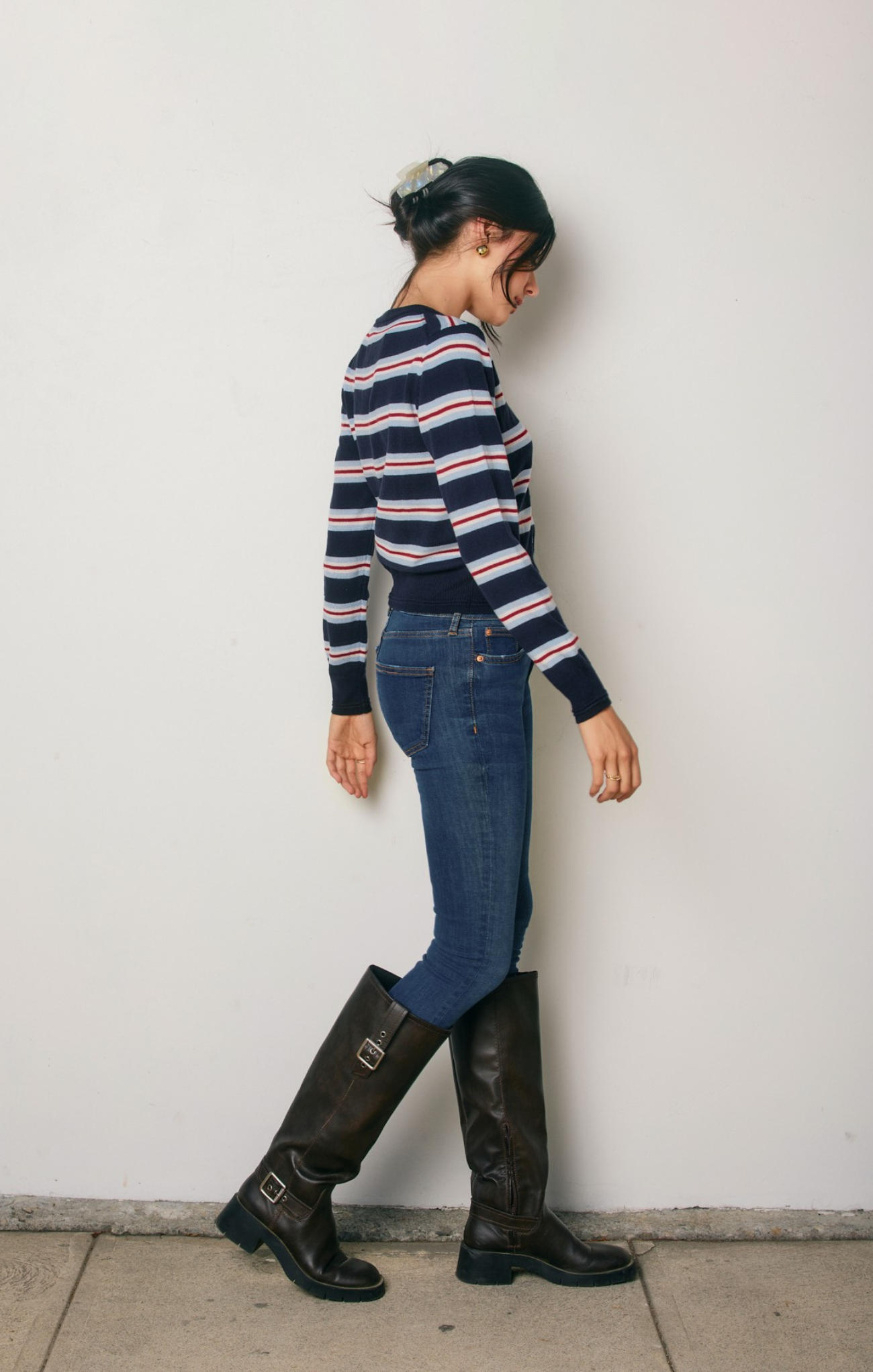 Person wearing a striped sweater, jeans, and knee-high boots against a white wall.