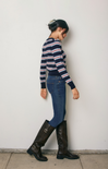 Person wearing a striped sweater, jeans, and knee-high boots against a white wall.