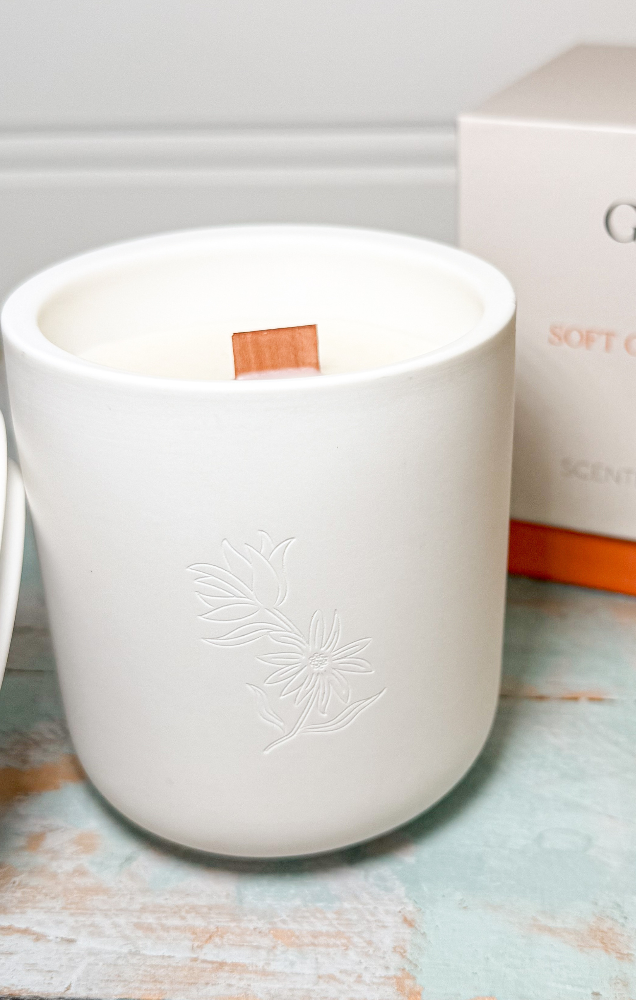 White candle in a decorative container with a box labeled 'SCENTED CANDLES' in the background.