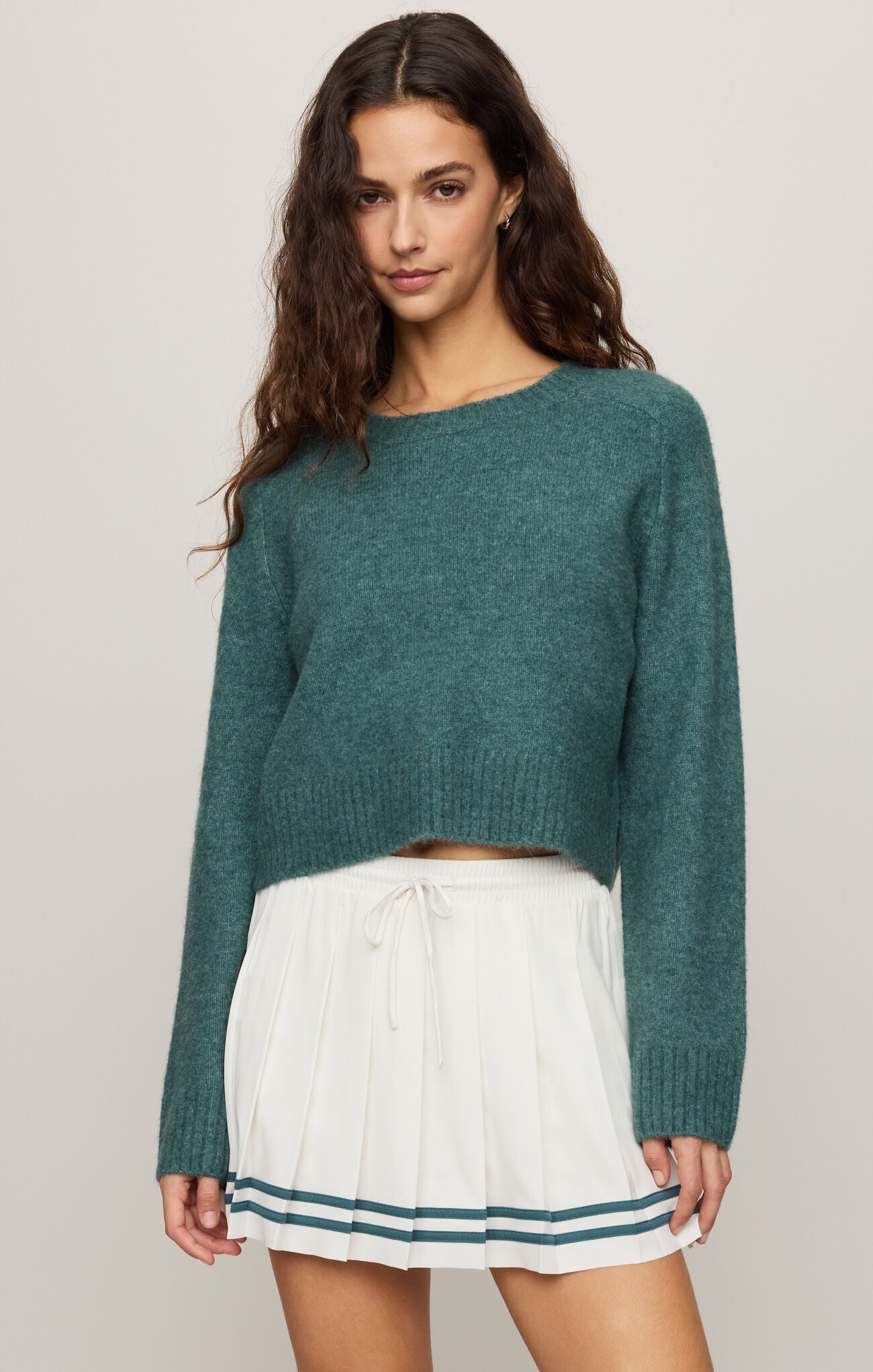 Woman wearing a teal sweater and white skirt with a striped hem on a plain background