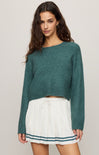 Woman wearing a teal sweater and white skirt with a striped hem on a plain background