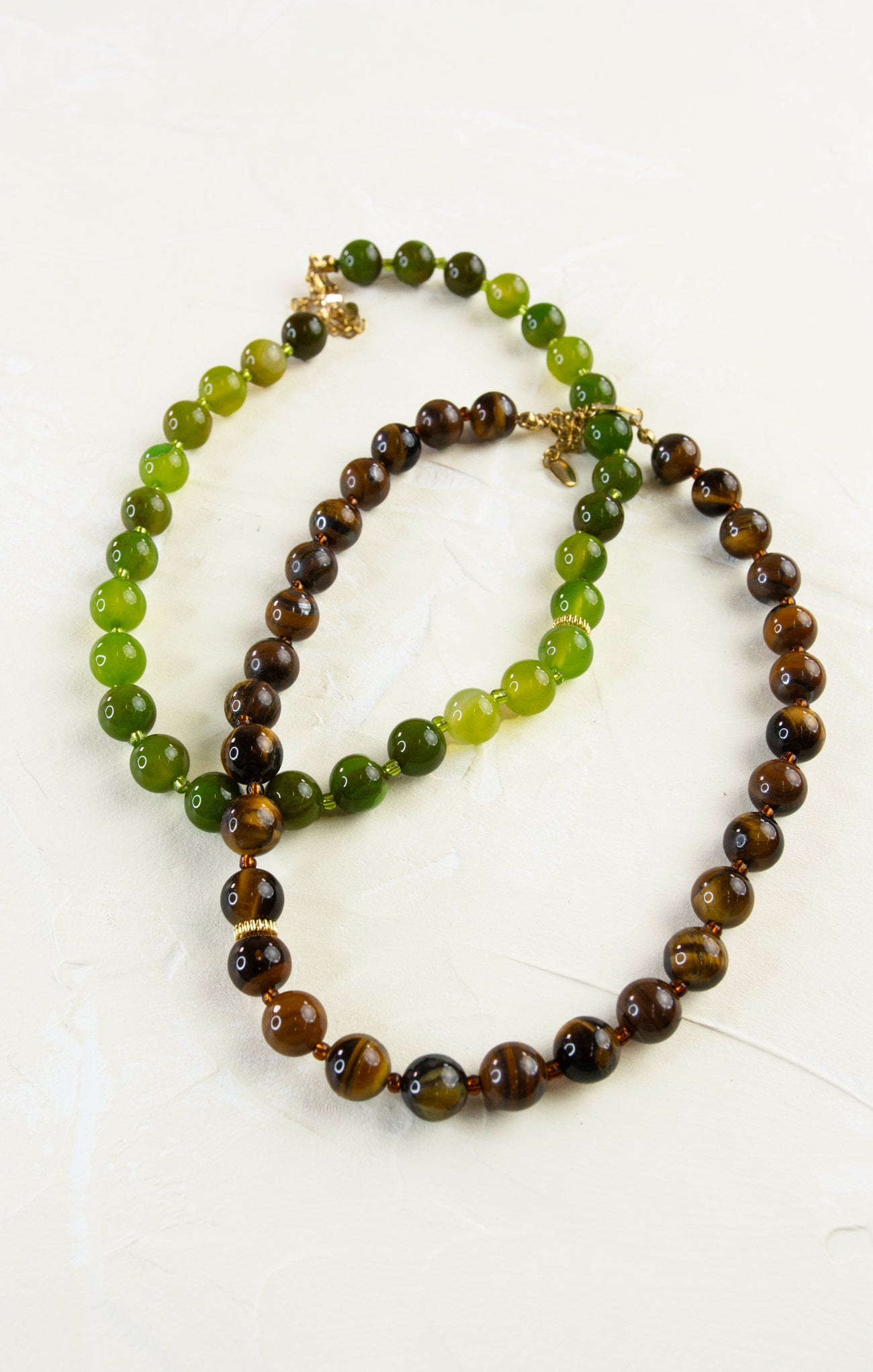 Two beaded necklaces, one green and brown, the other brown and black, on a white background.
