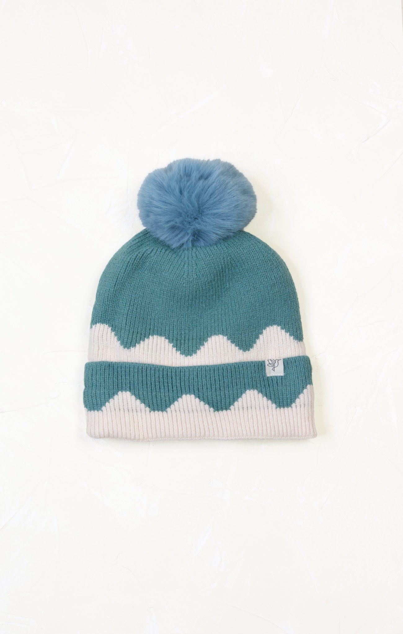 Teal knit beanie with white zigzag pattern and blue pom-pom on a white background