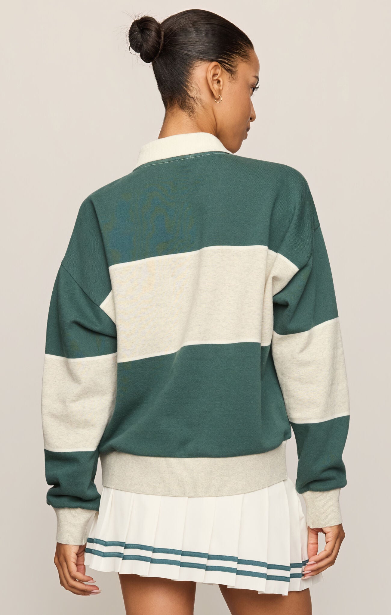 A woman shown from the back wearing a green and cream color-blocked knit sweater and a pleated white skirt with teal stripes, against a light beige studio background.