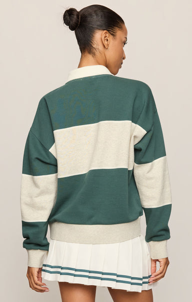 A woman shown from the back wearing a green and cream color-blocked knit sweater and a pleated white skirt with teal stripes, against a light beige studio background.