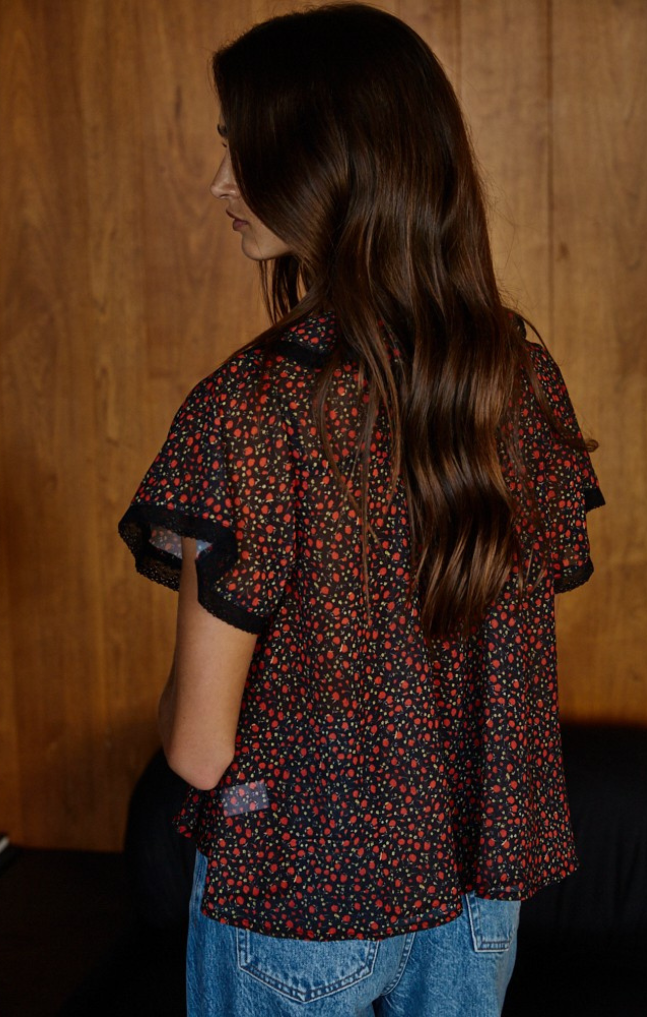 Person wearing a patterned blouse with short sleeves and blue jeans against a wooden wall.