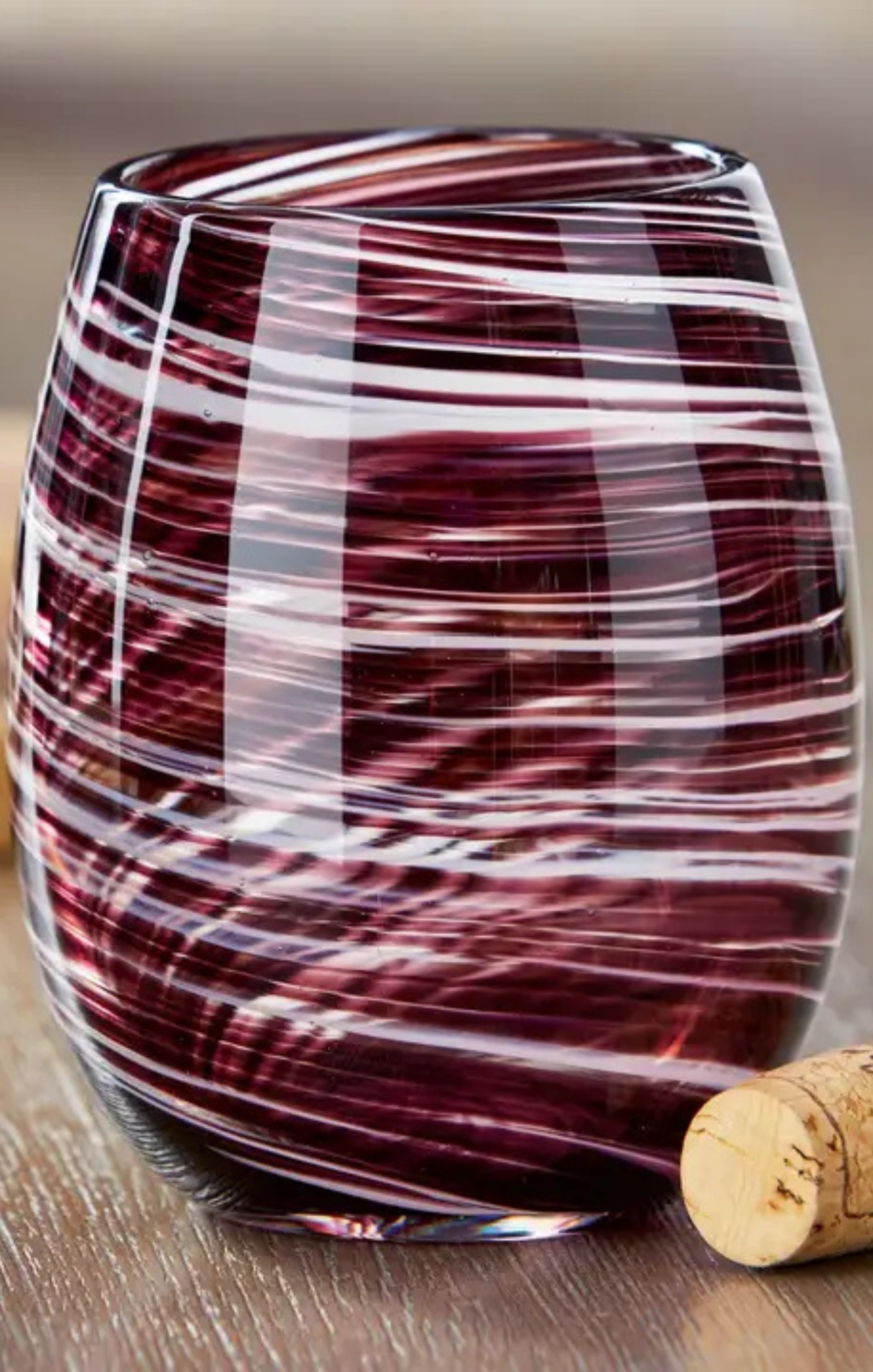 A stemless wine glass with a marbled pattern of deep burgundy and white swirls, shown on a wooden surface next to a cork.