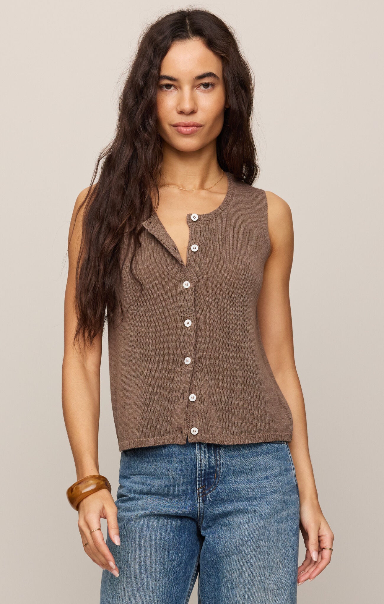 Woman wearing a brown sleeveless button-up top and blue jeans against a plain background
