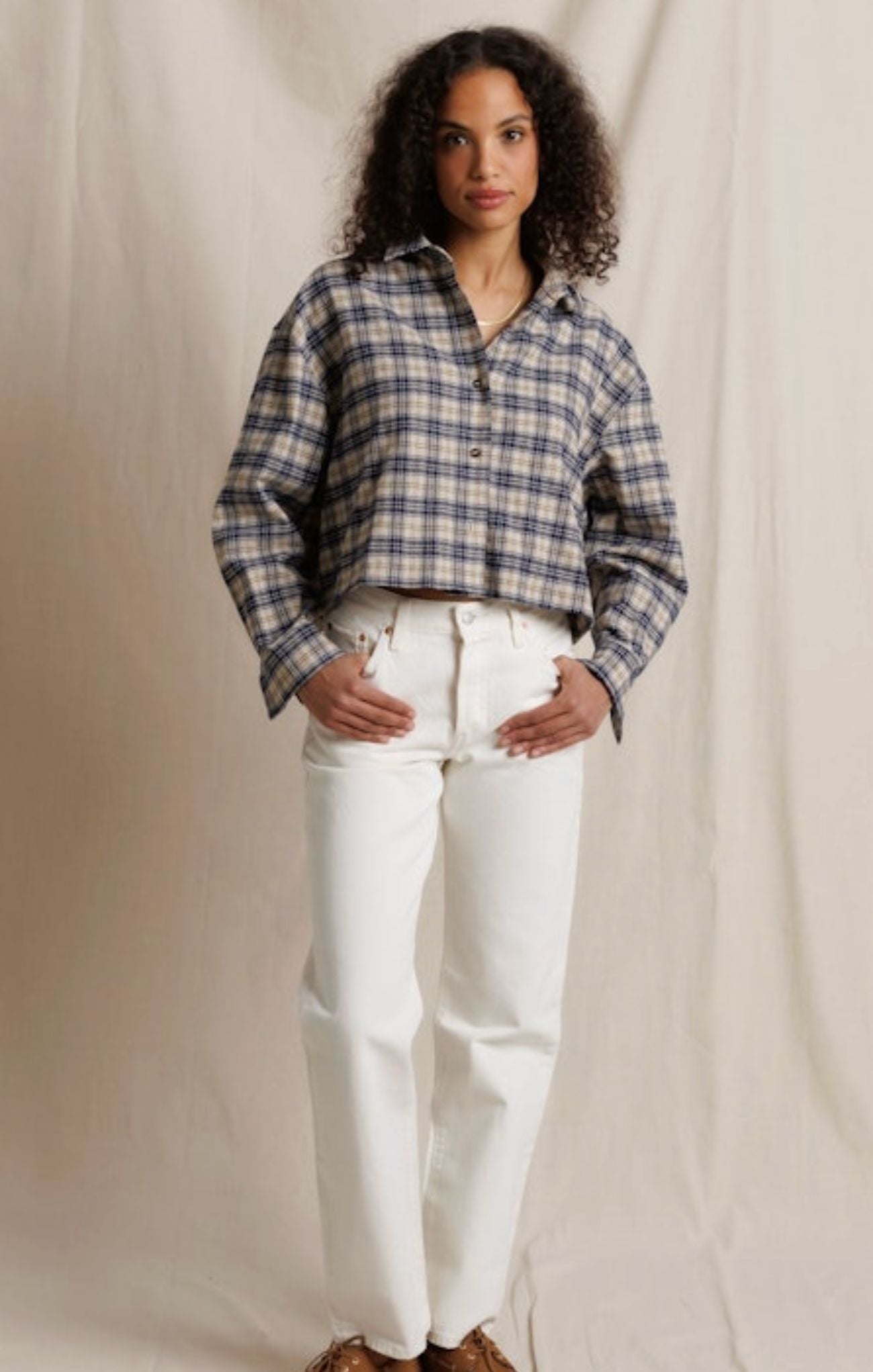 A woman stands against a draped beige fabric backdrop wearing a cropped, long-sleeve plaid button-up shirt in blue, white, and gray, paired with white pants and brown shoes.