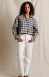 A woman stands against a draped beige fabric backdrop wearing a cropped, long-sleeve plaid button-up shirt in blue, white, and gray, paired with white pants and brown shoes.