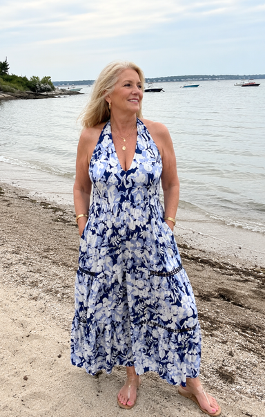 Woman in a blue floral dress standing on a beach with water and boats in the background