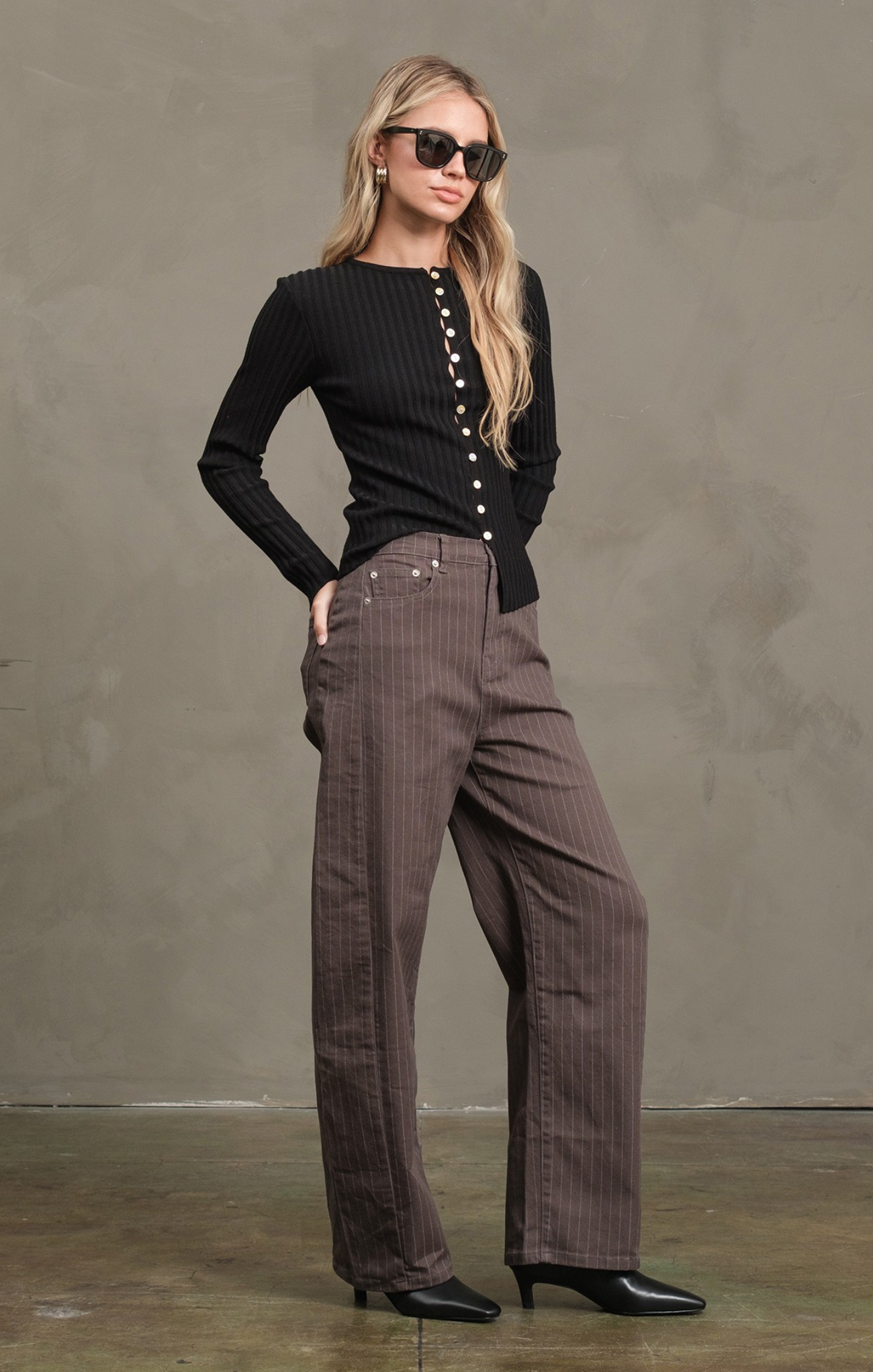 Woman wearing a black ribbed sweater and brown plaid pants against a gray background