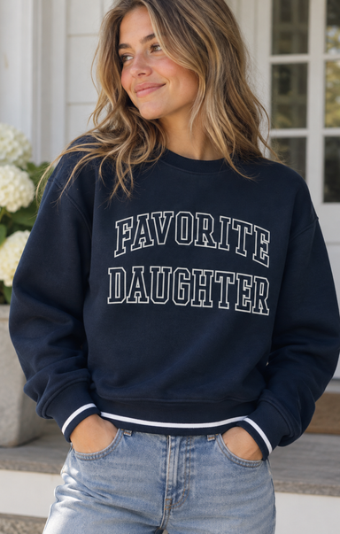 A woman wearing a navy blue crewneck sweatshirt with 'FAVORITE DAUGHTER' printed in white varsity-style letters across the chest, paired with light-wash jeans.
