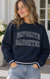 A woman wearing a navy blue crewneck sweatshirt with 'FAVORITE DAUGHTER' printed in white varsity-style letters across the chest, paired with light-wash jeans.