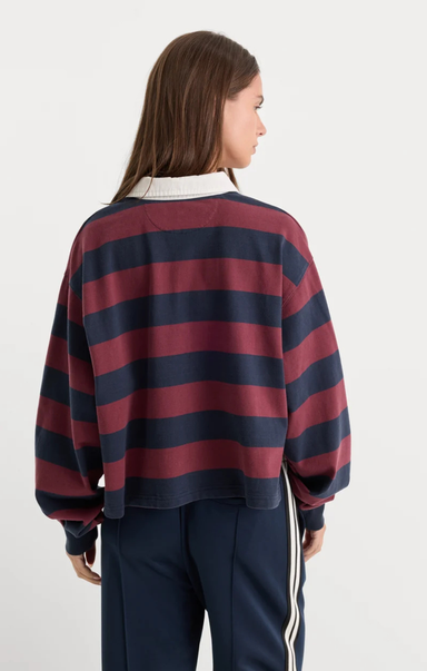 A woman shown from the back wearing a cropped long-sleeve rugby-style top with horizontal navy and burgundy stripes, paired with navy track pants with white side stripes, against a plain light gray studio background.