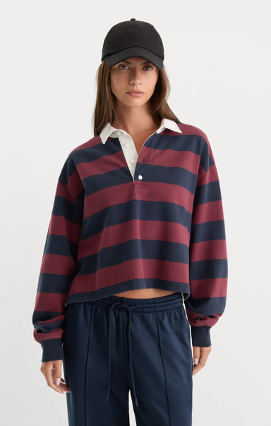 A woman modeling a cropped long-sleeve rugby shirt with horizontal navy and burgundy stripes, layered over a white collared shirt, paired with navy drawstring sweatpants and a black baseball cap, against a light gray studio background.
