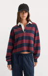 A woman modeling a cropped long-sleeve rugby shirt with horizontal navy and burgundy stripes, layered over a white collared shirt, paired with navy drawstring sweatpants and a black baseball cap, against a light gray studio background.