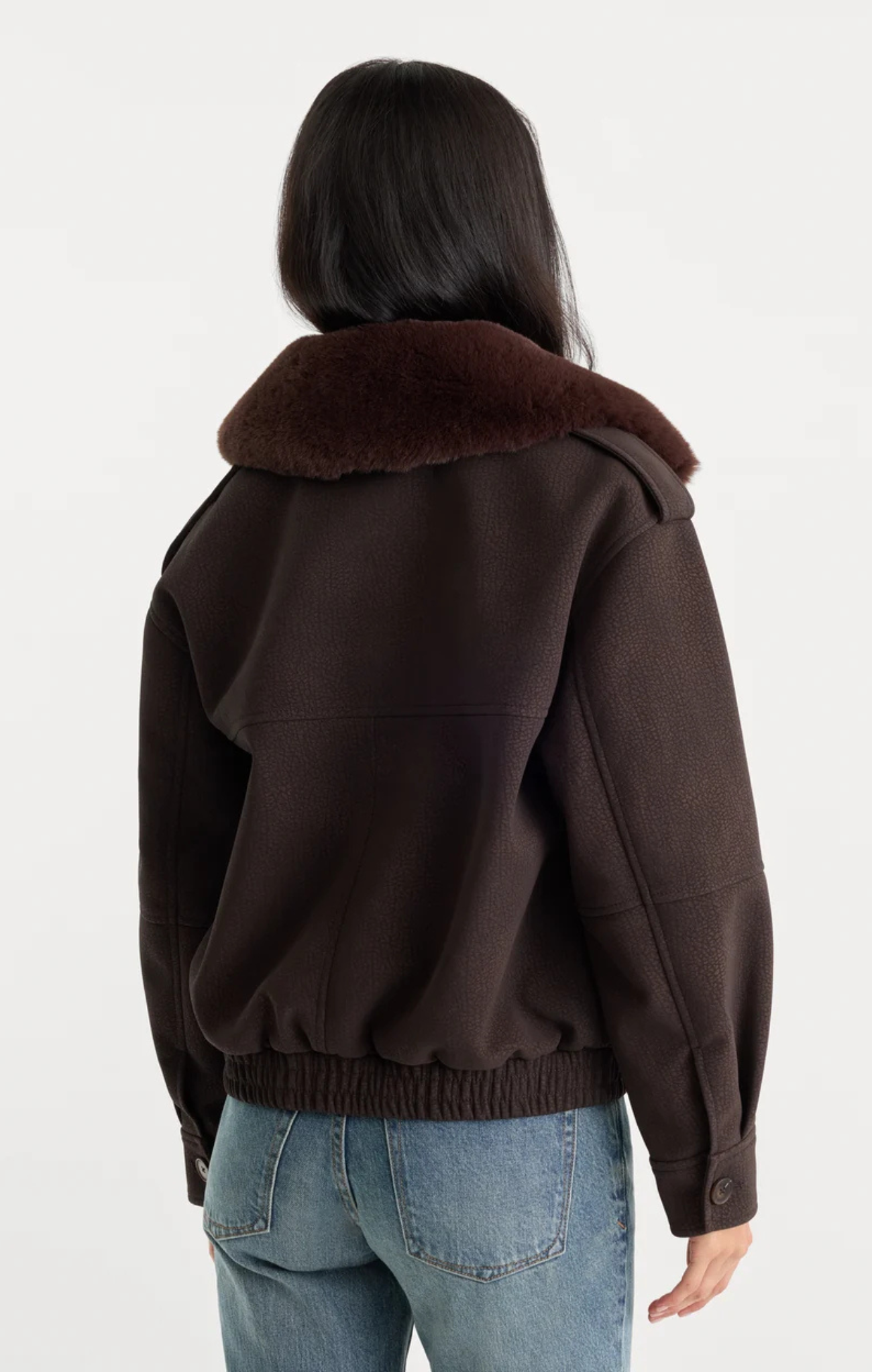 A woman viewed from the back wearing a dark brown jacket with a thick dark brown shearling-style collar and light blue jeans, against a plain white studio background.