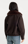 A woman viewed from the back wearing a dark brown jacket with a thick dark brown shearling-style collar and light blue jeans, against a plain white studio background.
