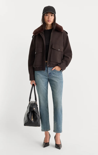 A model stands against a light gray studio background wearing a dark brown shearling-lined aviator jacket over a black top, blue jeans, black heels, and a black cap, while holding a black handbag.