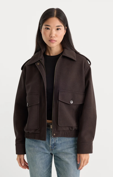 A woman modeling a dark brown cropped jacket with flap pockets over a black top and blue jeans, photographed against a light gray studio background.