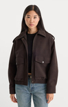 A woman modeling a dark brown cropped jacket with flap pockets over a black top and blue jeans, photographed against a light gray studio background.