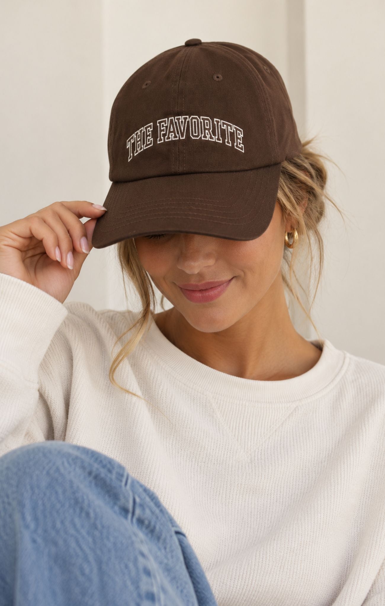 A woman wearing a brown baseball cap with 'THE FAVORITE' embroidered in white arched lettering, posing in a white ribbed sweatshirt and blue jeans against a neutral indoor background.
