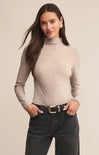 Woman wearing a beige turtleneck sweater and dark jeans on a plain background