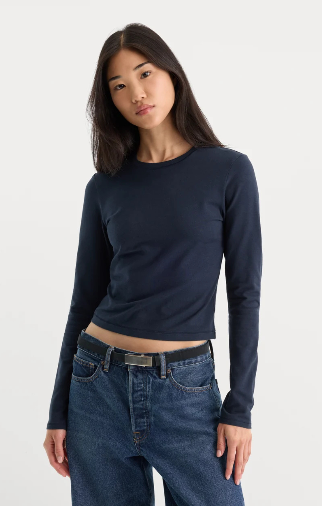 A woman modeling a dark navy blue long-sleeve crop top paired with blue jeans against a plain light gray studio background.