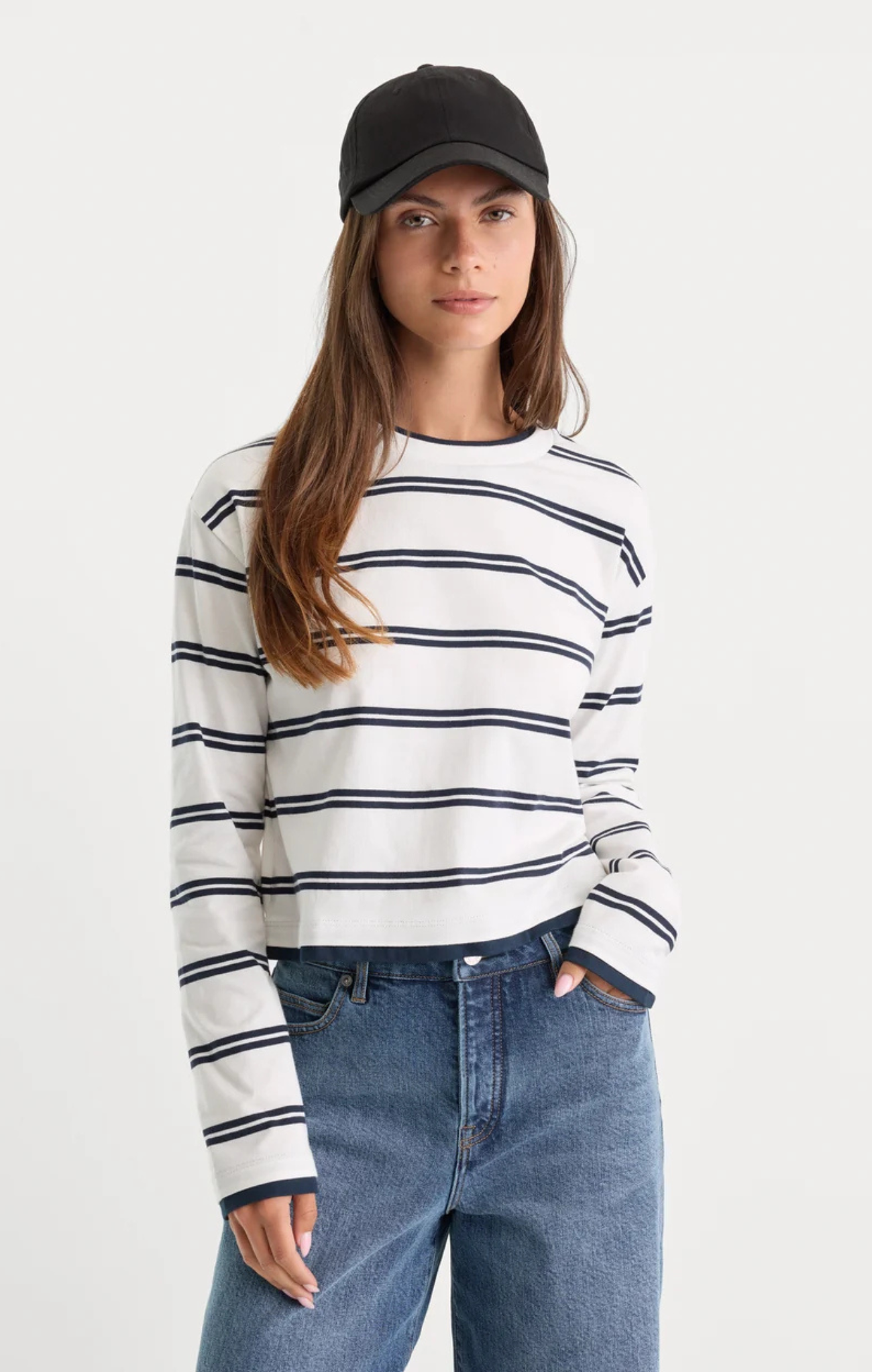 A woman wearing a white long-sleeve crewneck sweater with navy blue horizontal stripes, paired with blue jeans and a black baseball cap, photographed against a light gray studio background.