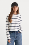 A woman wearing a white long-sleeve crewneck sweater with navy blue horizontal stripes, paired with blue jeans and a black baseball cap, photographed against a light gray studio background.