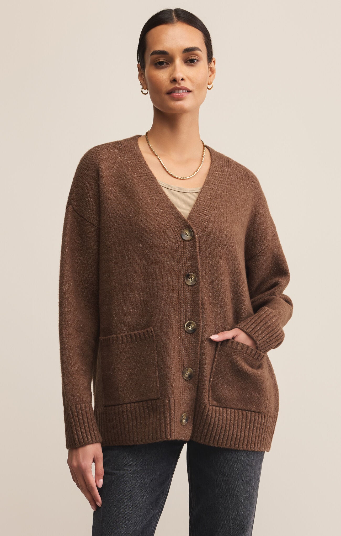 A female model wearing a brown cardigan with buttons, V-neckline, and ribbed hem, paired with a white top and blue jeans.