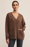A female model wearing a brown cardigan with buttons, V-neckline, and ribbed hem, paired with a white top and blue jeans.