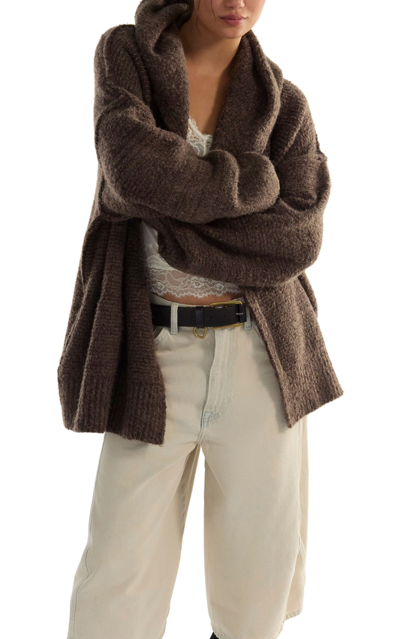 A person wearing a chunky, oversized brown knit cardigan or wrap sweater over a white lace top and cream wide-leg pants, photographed against a plain white studio background.