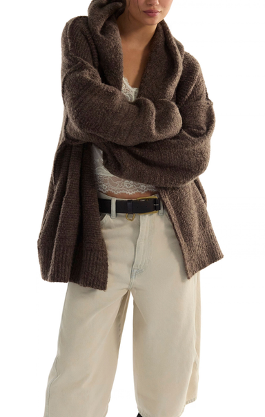 A person wearing a chunky, oversized brown knit cardigan or wrap sweater over a white lace top and cream wide-leg pants, photographed against a plain white studio background.