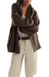 A person wearing a chunky, oversized brown knit cardigan or wrap sweater over a white lace top and cream wide-leg pants, photographed against a plain white studio background.