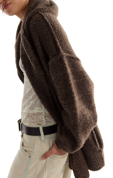 A close-up side view of a person wearing a chunky, oversized brown fuzzy cardigan or shrug over a cream lace top and light beige jeans with a black belt, against a plain white background.