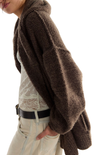 A close-up side view of a person wearing a chunky, oversized brown fuzzy cardigan or shrug over a cream lace top and light beige jeans with a black belt, against a plain white background.