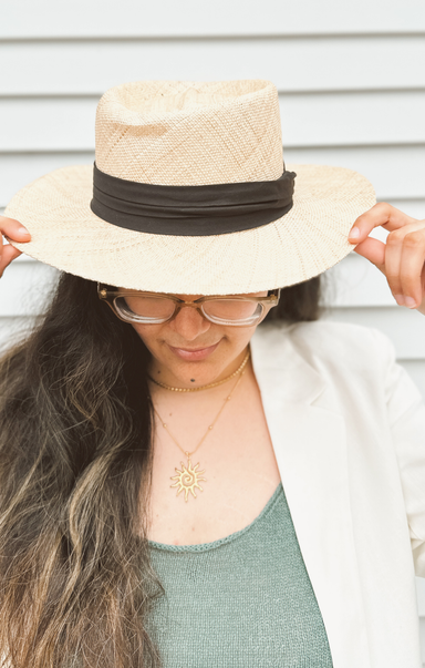 Woman wearing a straw hat with a black band, glasses, and a necklace with a sun pendant.