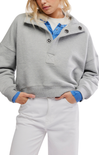 A model wearing a light gray heathered cropped sweatshirt with a mock neck and decorative buttons, layered over a blue collared shirt and white pants.
