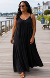 Woman wearing a black dress standing on a wooden boardwalk by a waterfront.