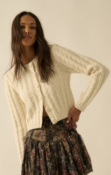 Woman wearing a cream cable knit cardigan over a patterned skirt on a plain background